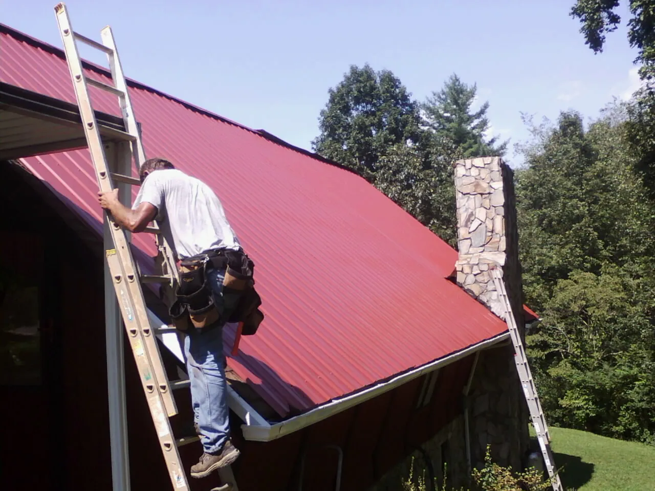 Roofer on a ladder beside a red metal roof and stone chimney — Soffit & Fascia Repair by Roofing Brothers in Burkeville